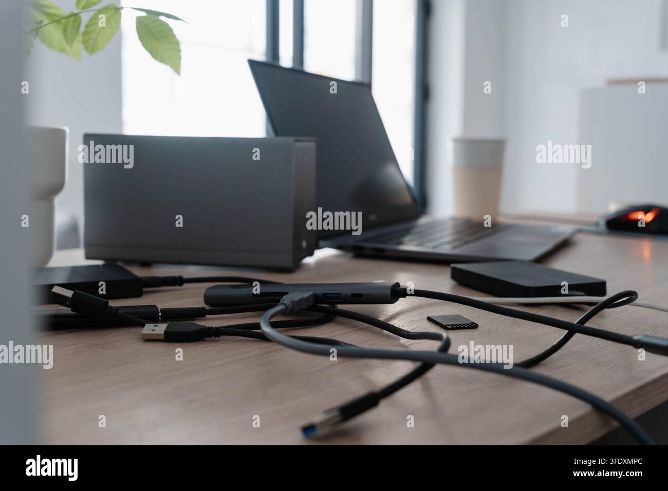 Cavi USB ingarbugliati, un hub e dischi rigidi portatili ingombrano una scrivania di legno accanto a un laptop e un monitor esterno. Lo spazio di archiviazione cloud sostituisce le connessioni cablate disordinate Foto Stock