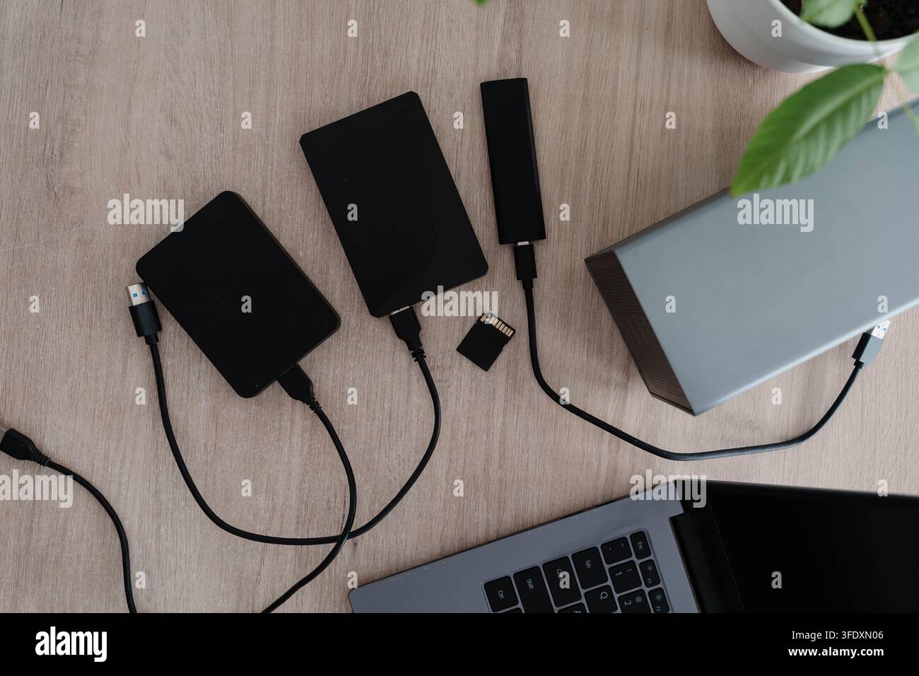 Dischi rigidi esterni, un SSD portatile e una scheda di memoria giacciono con cavi su una scrivania di legno accanto a un laptop aperto. Configurazione di archiviazione e backup dei dati per Foto Stock