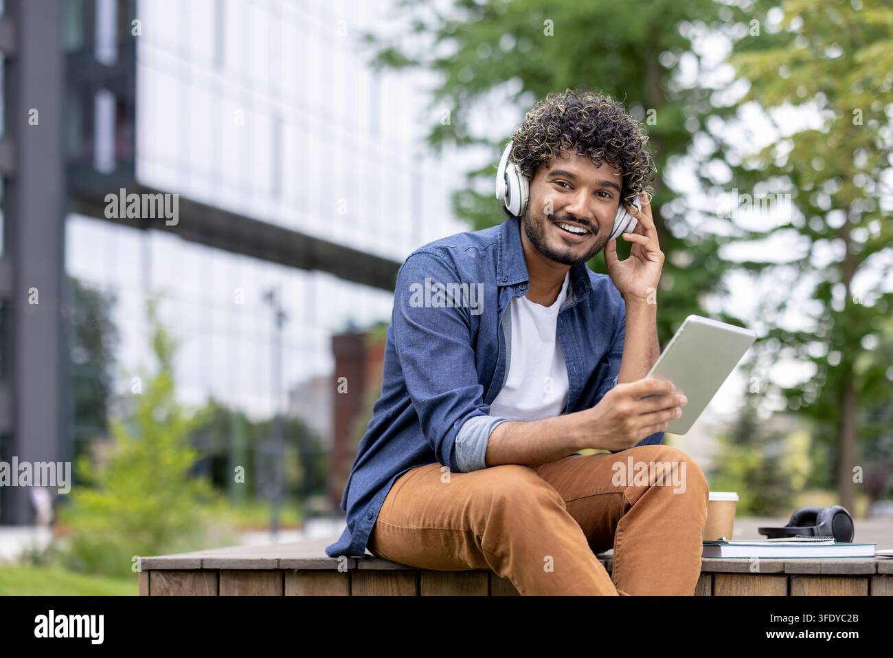 Giovane studente felice con i capelli ricci che indossa cuffie, sorride e tiene un tablet digitale seduto su una panchina di legno all'aperto vicino a un edificio moderno del campus universitario Foto Stock