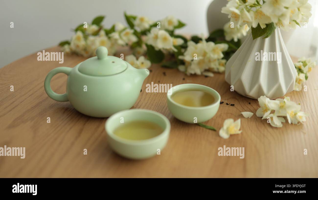 Servizio da tè verde tradizionale con teiera in ceramica, piccole tazze e vaso con fiori bianchi su tavolo di legno, composizione pacifica di natura morta. Foto Stock