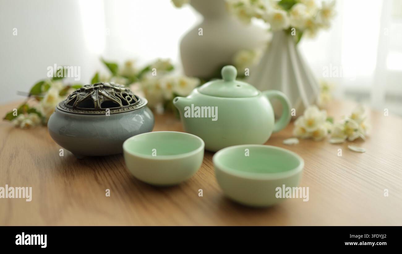 Servizio da tè verde tradizionale con teiera in ceramica, piccole tazze e vaso con fiori bianchi su tavolo di legno, composizione pacifica di natura morta. Foto Stock