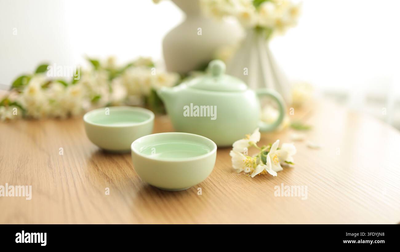Servizio da tè verde tradizionale con teiera in ceramica, piccole tazze e vaso con fiori bianchi su tavolo di legno, composizione pacifica di natura morta. Foto Stock