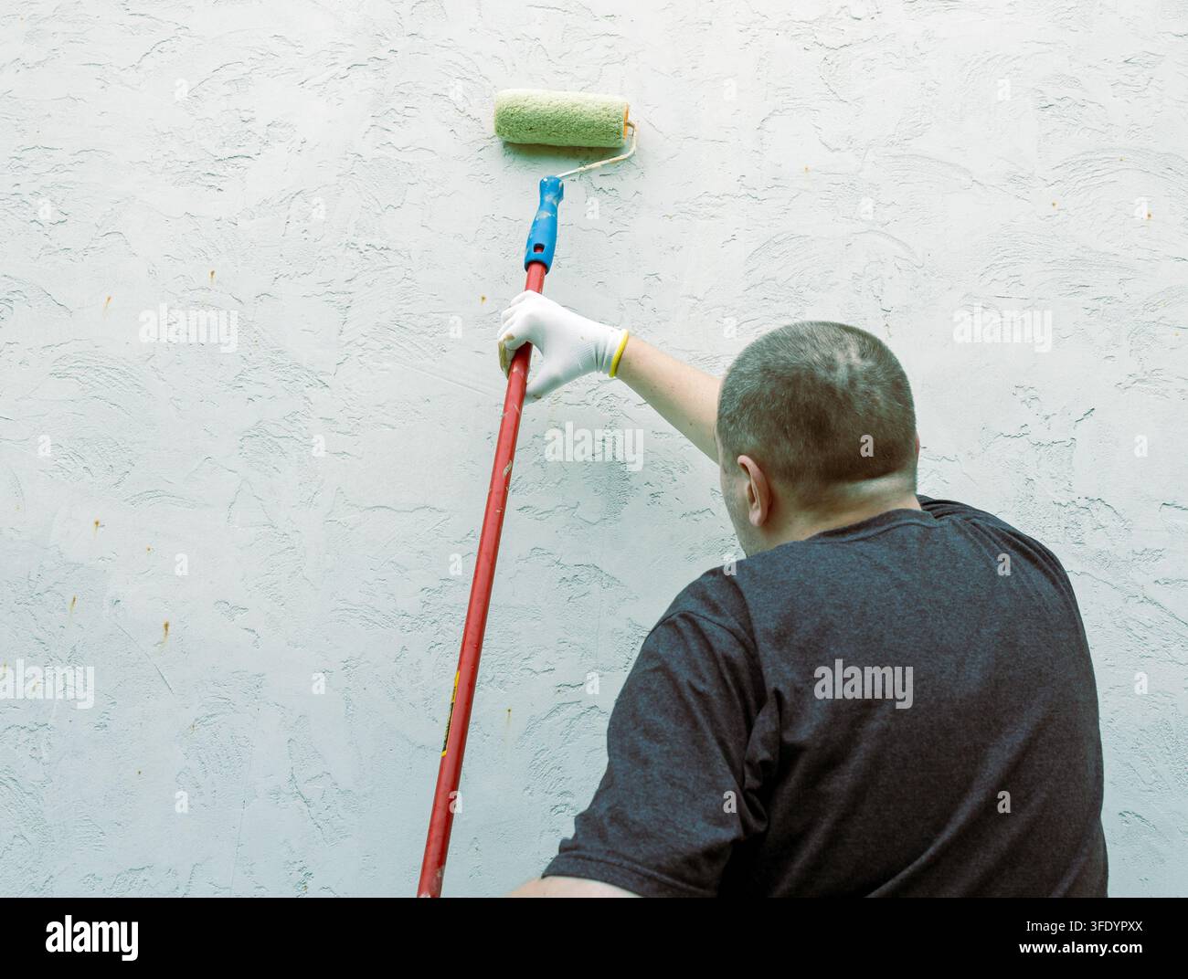 Uomo rotola la vernice su un muro ruvido. Mano guantata afferra un rullo a manico lungo Foto Stock