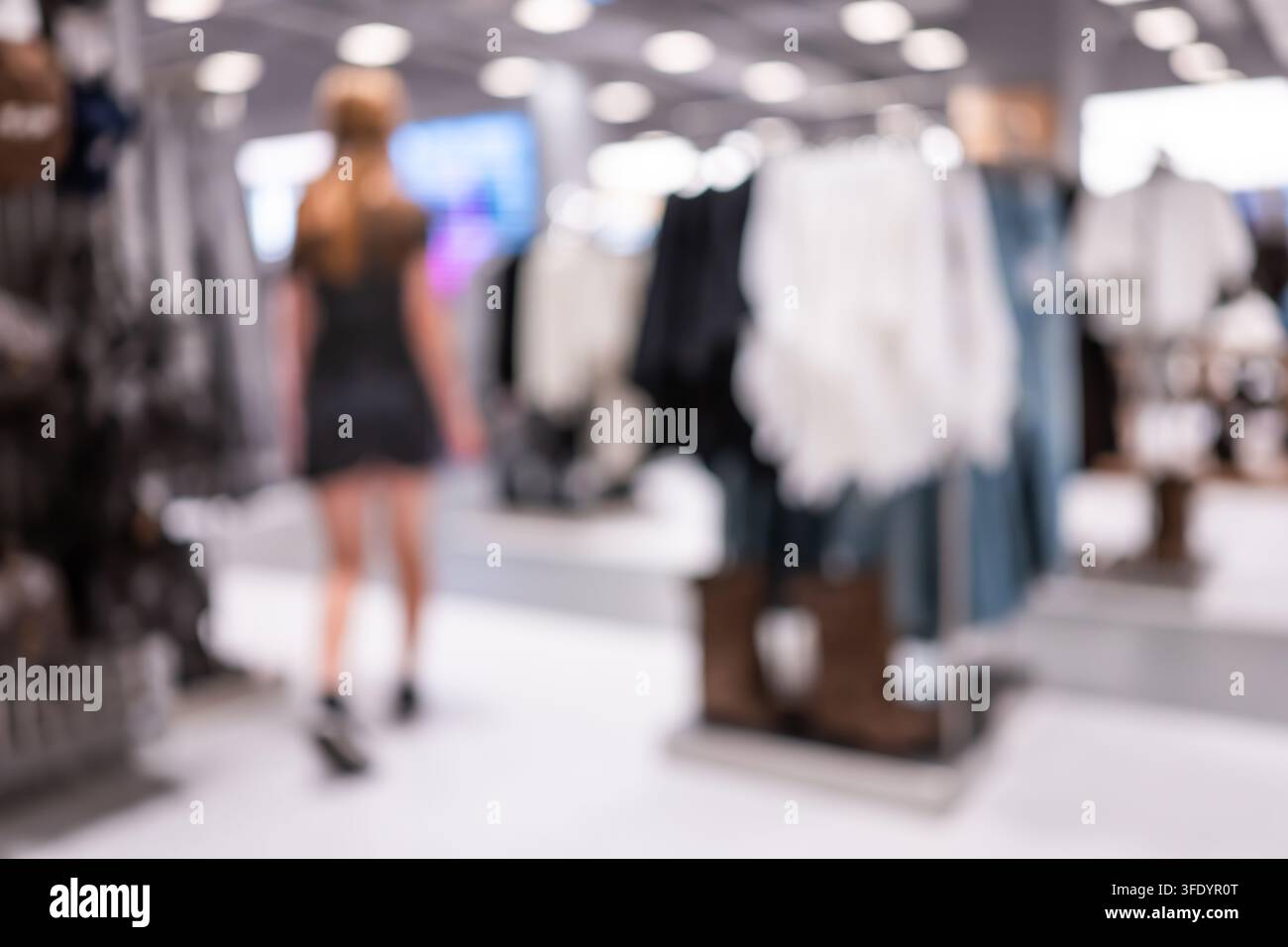 Donna sfocata che cammina tra i corridoi di abiti alla moda in un negozio al dettaglio moderno e luminoso Foto Stock
