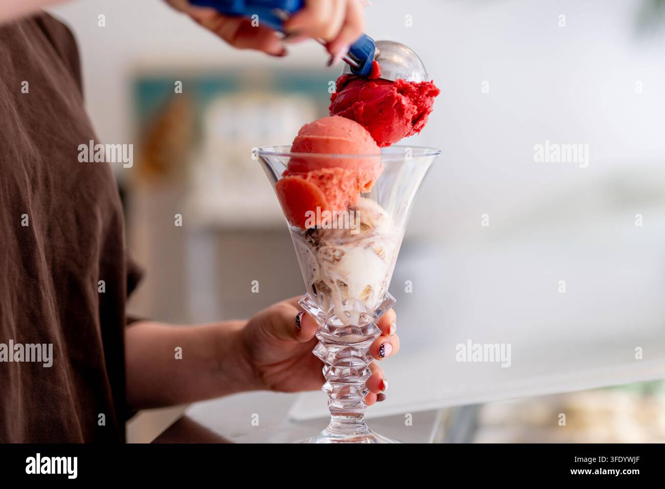 Una persona aggiunge una pallina di gelato rosso ricco a un bicchiere da sundae trasparente contenente aromi di pesca e noci. Foto Stock