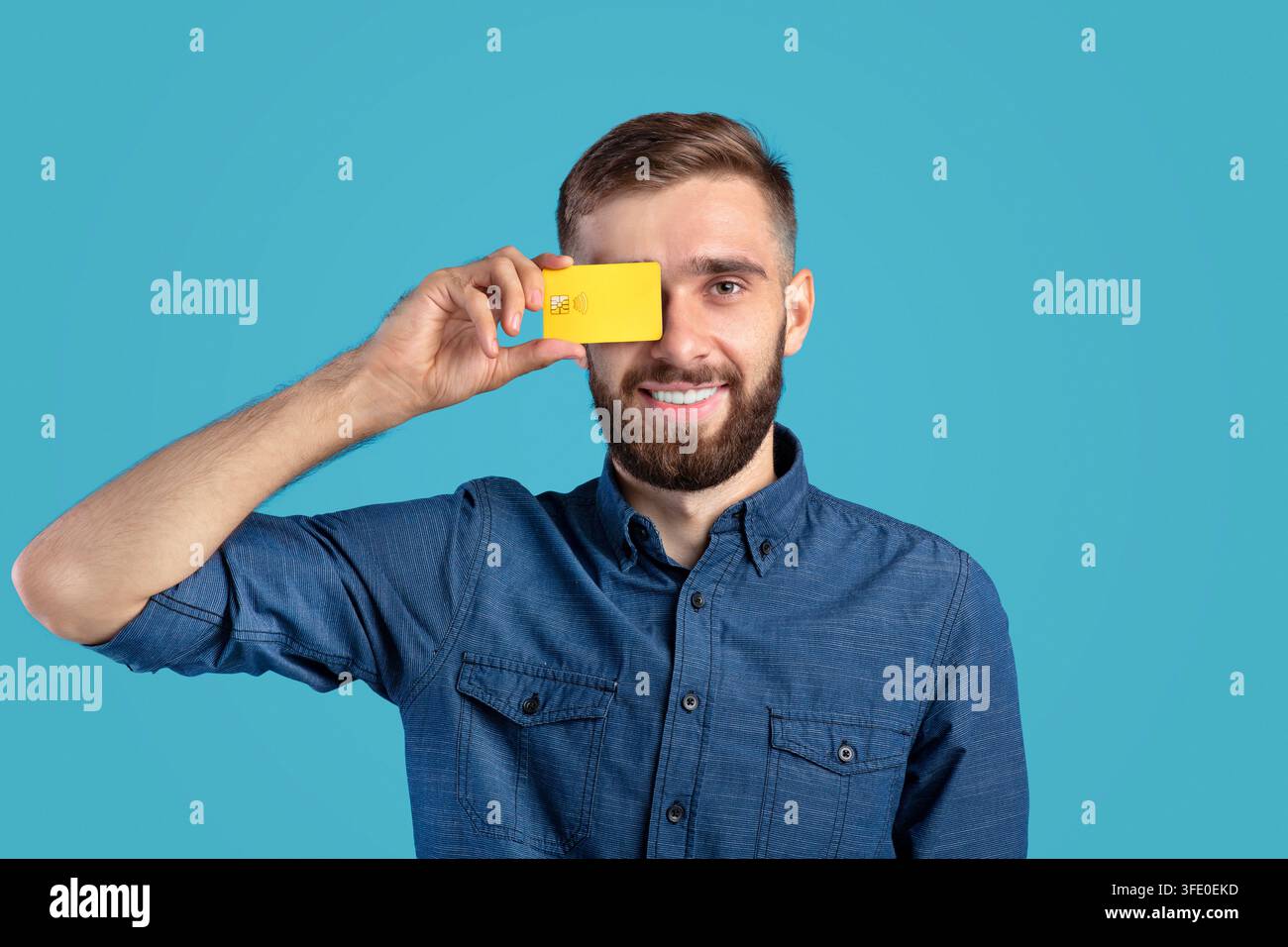 Ragazzo milleniale sorridente che tiene una carta di credito davanti all'occhio su sfondo blu Foto Stock