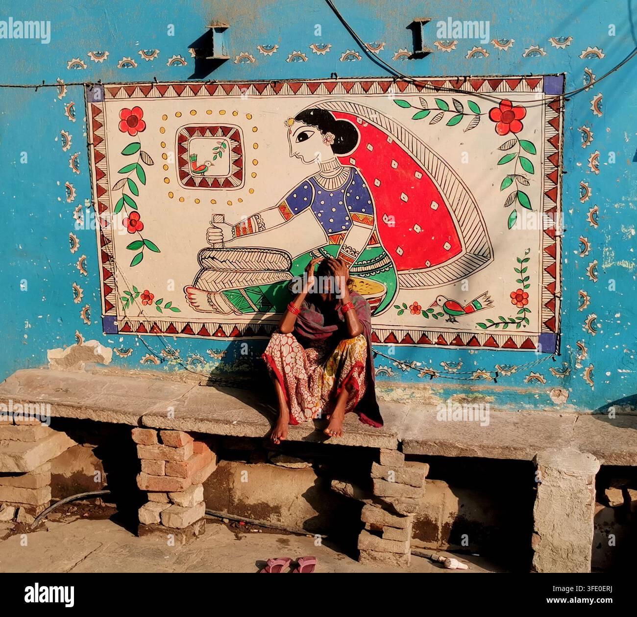 VARANASI, INDIA - 23 GENNAIO 2026: Donna indiana seduta davanti a un colorato dipinto murale tradizionale su un muro di strada, Varanasi, India Foto Stock