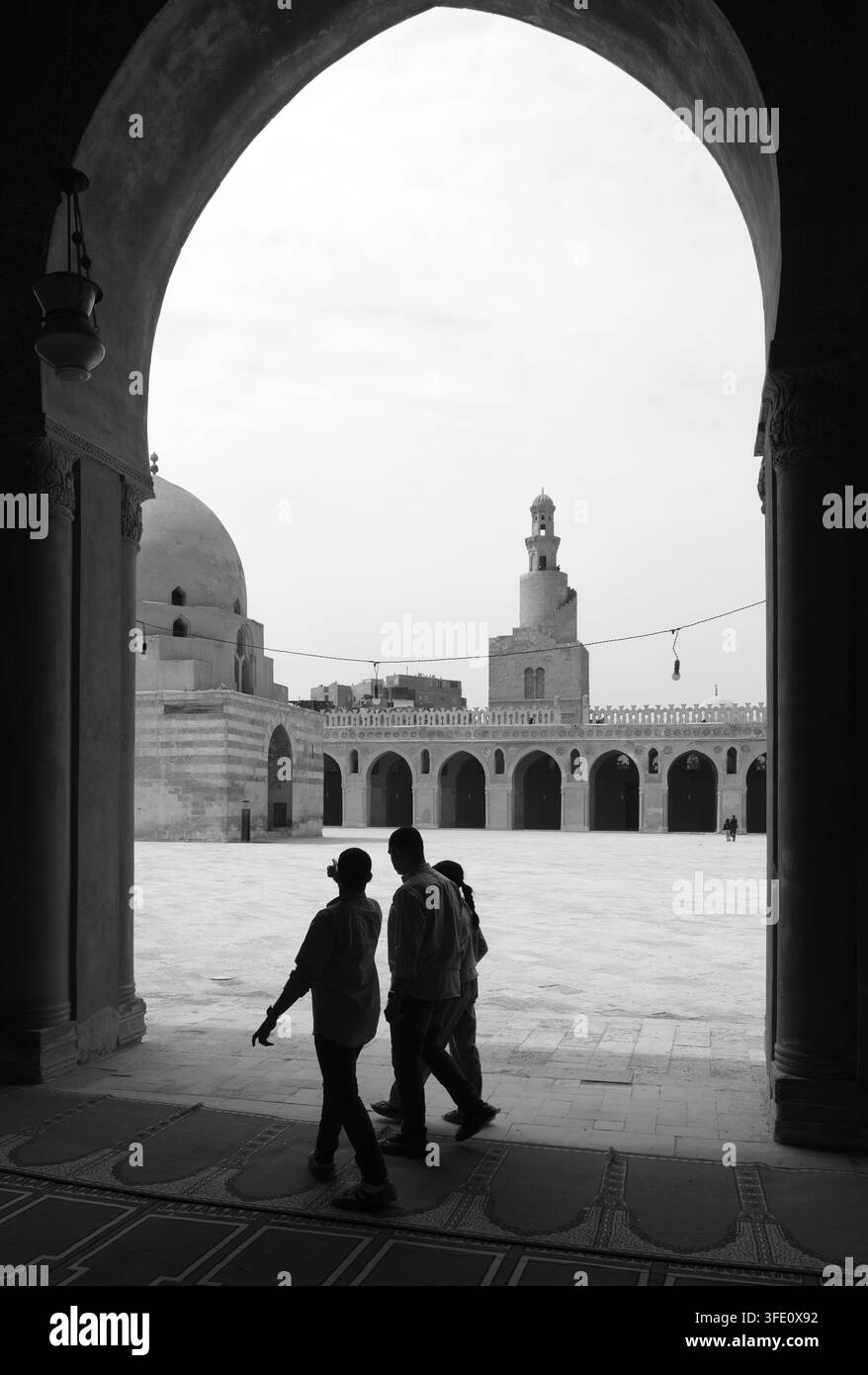 CAIRO, EGITTO: Visitatori in controluce camminano da un arco buio verso il cortile illuminato della moschea di Ibn Tulun, rivelando il suo minareto a spirale Foto Stock