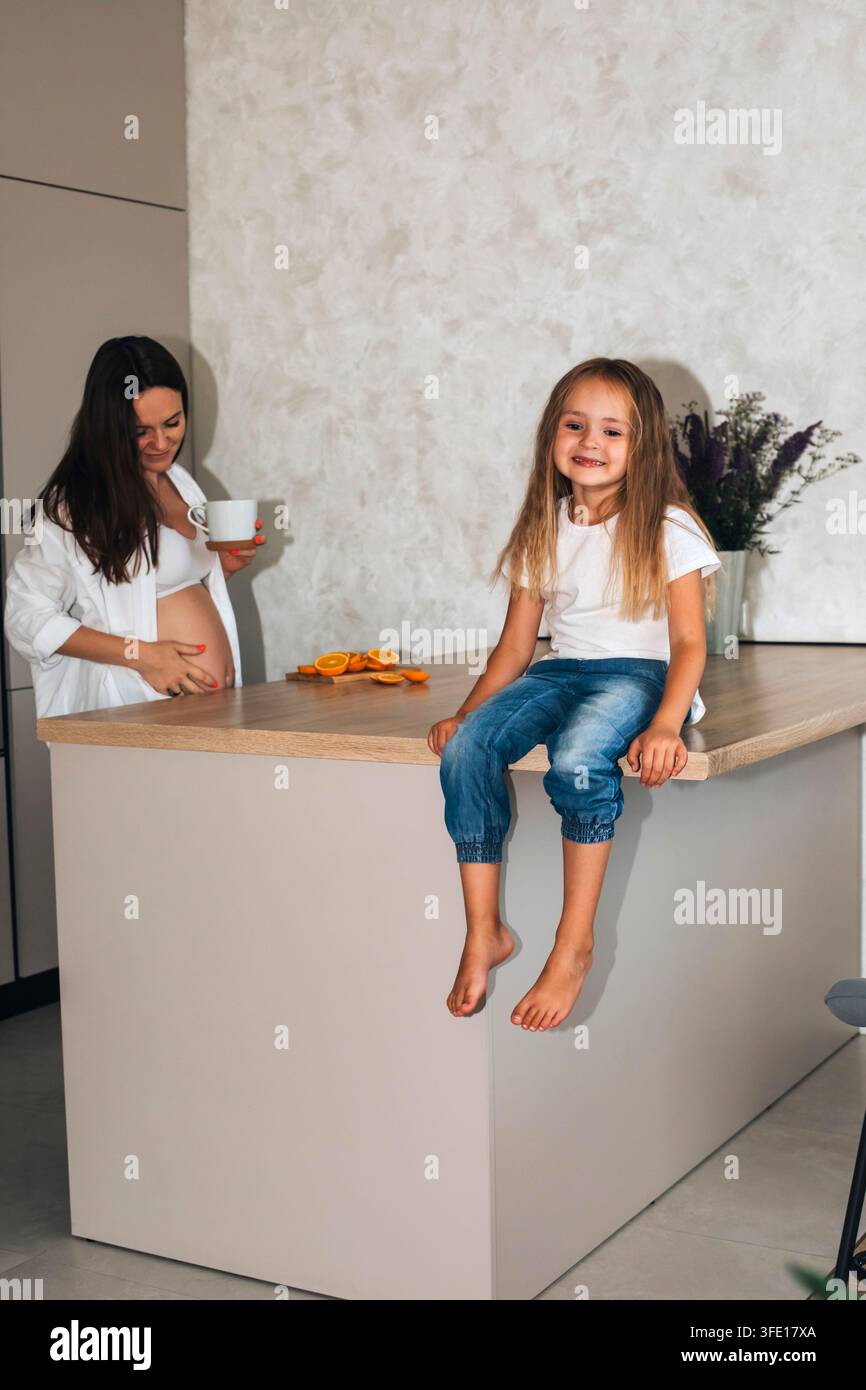 Madre incinta e figlia felice che passano del tempo insieme in una cucina moderna Foto Stock