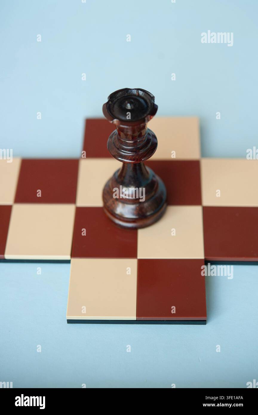 Primo piano di una regina degli scacchi in legno scuro in piedi su una piccola scacchiera rossa e crema su uno sfondo blu pallido Foto Stock