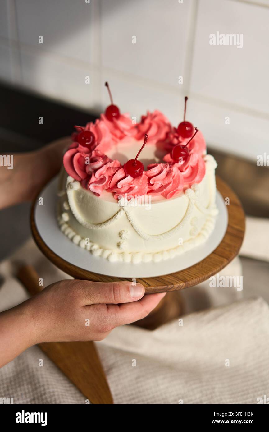 Mani che tengono una piccola torta bianca con rosette di glassa rosa e ciliegie, con dettagli di torta, mano, glassa, ciliegia, che mostrano crema al burro, rosetta, supporto Foto Stock