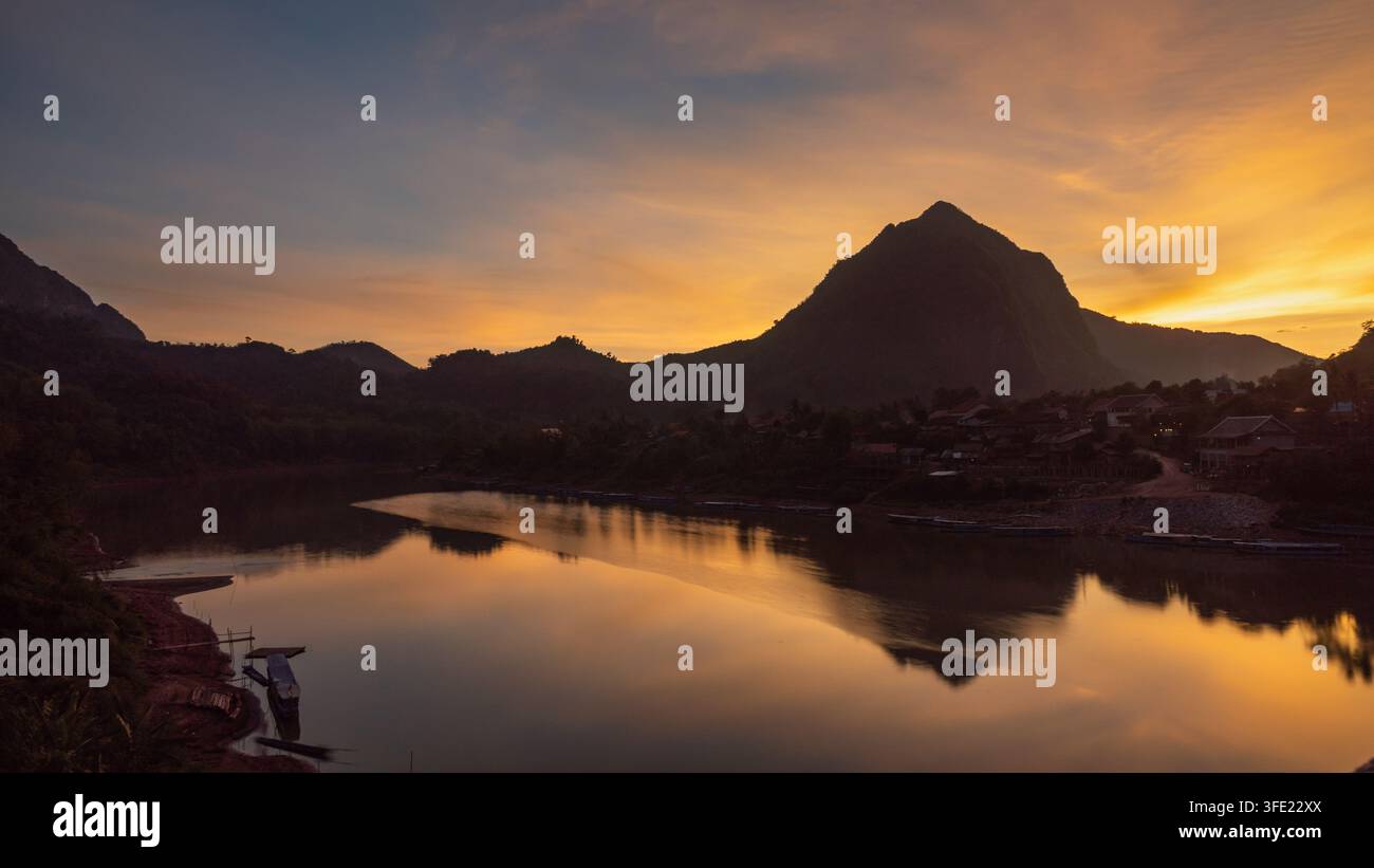 Vista aerea del fiume Nam Ou che scorre davanti alle case del villaggio al tramonto con silhouette di montagne e luce dorata che si riflette sull'acqua a Nong Khiaw Foto Stock