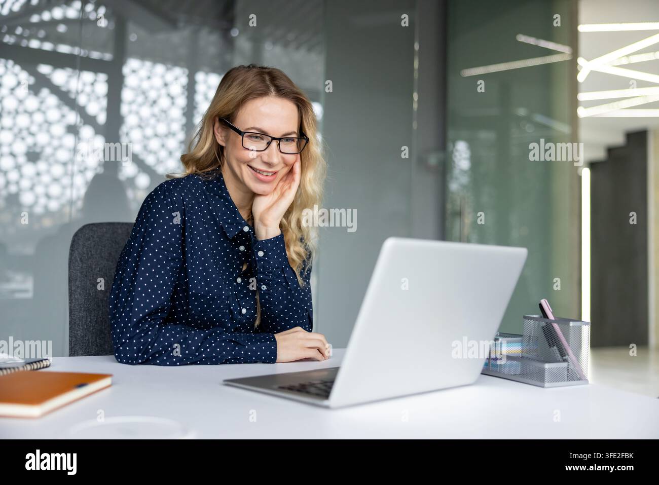 Donna d'affari sorridente con occhiali che collabora da remoto e interagisce con contenuti digitali sul suo laptop, incarnando un ambiente di lavoro professionale e positivo mentre utilizza la tecnologia Foto Stock