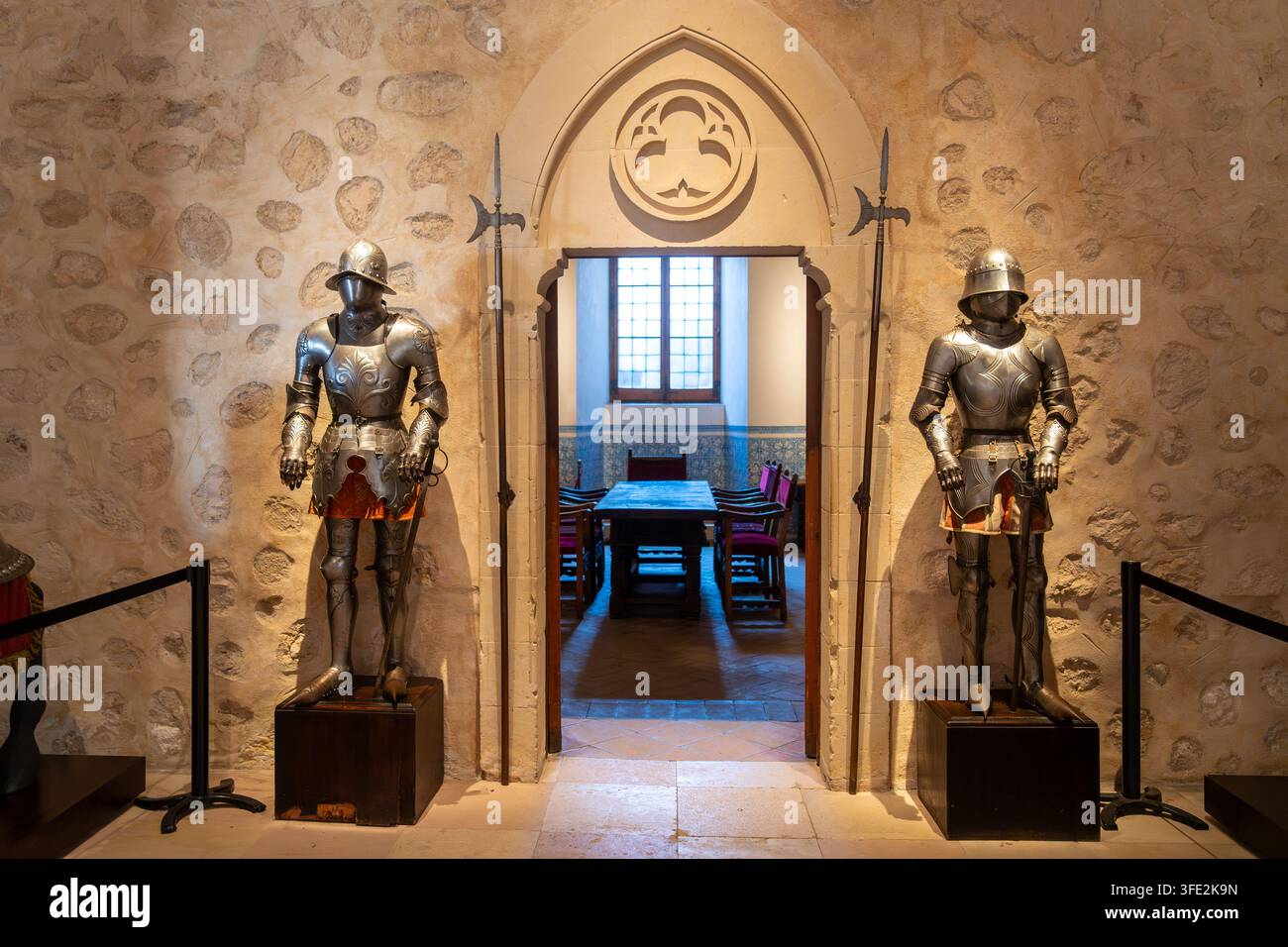 Due complete armature da cavaliere fanno la guardia accanto a un portale in pietra ad arco che conduce a una sala da pranzo illuminata dal sole nell'interno di un castello storico. Luce calda Foto Stock