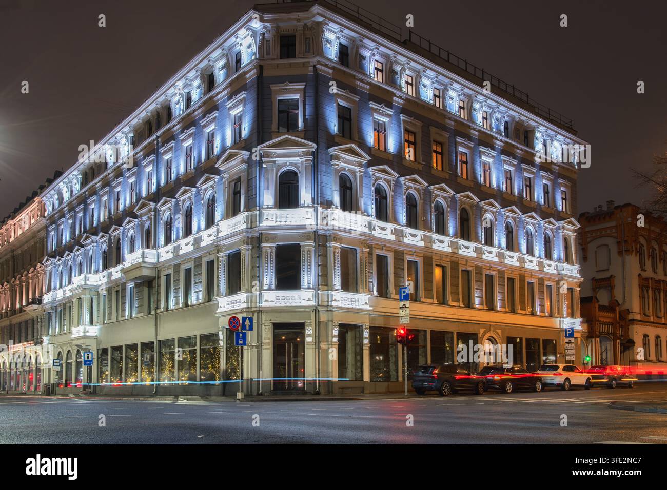Facciata luminosa e traffico definiscono una veduta notturna verticale di Riga dove un edificio angolare ornato si erge sopra i veicoli in movimento, adatto all'architettura, cit Foto Stock