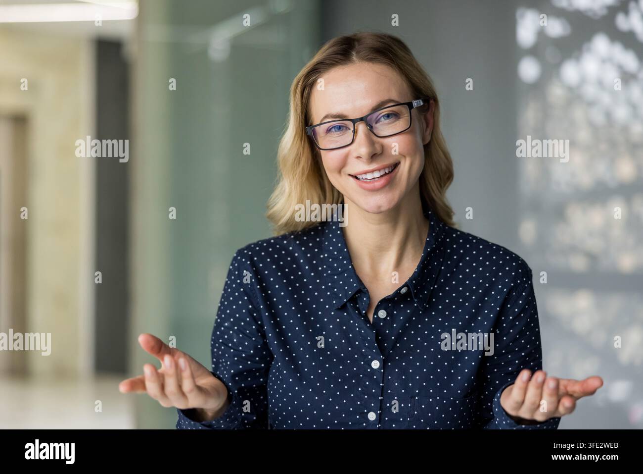 Donna d'affari bionda con occhiali e camicia a pois, guarda la telecamera con un sorriso accogliente e mani aperte, comunica sicurezza e cordialità in un ambiente d'ufficio Foto Stock