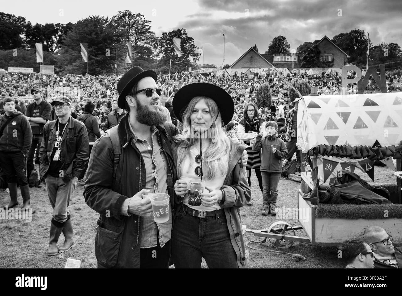 COPIA STILEVOLE AL FESTIVAL GREEN MAN, REPORTAGE, FESTIVAL GREEN MAN 2017, BRECON, WALES, UK, 2017: Una coppia giovane e stilosa con cappelli e occhiali da sole che tiene bicchieri di plastica con birra è in piedi insieme in un campo, con una grande folla del festival visibile sullo sfondo dietro di loro. Foto: Rob Watkins. INFO: Il Festival Green Man è iniziato nel 2003 in Galles. Si tiene ogni anno nelle Brecon Beacons il terzo fine settimana di agosto e celebra musica alternativa, arte, letteratura, scienza e cultura familiare in uno splendido scenario montano. Foto Stock