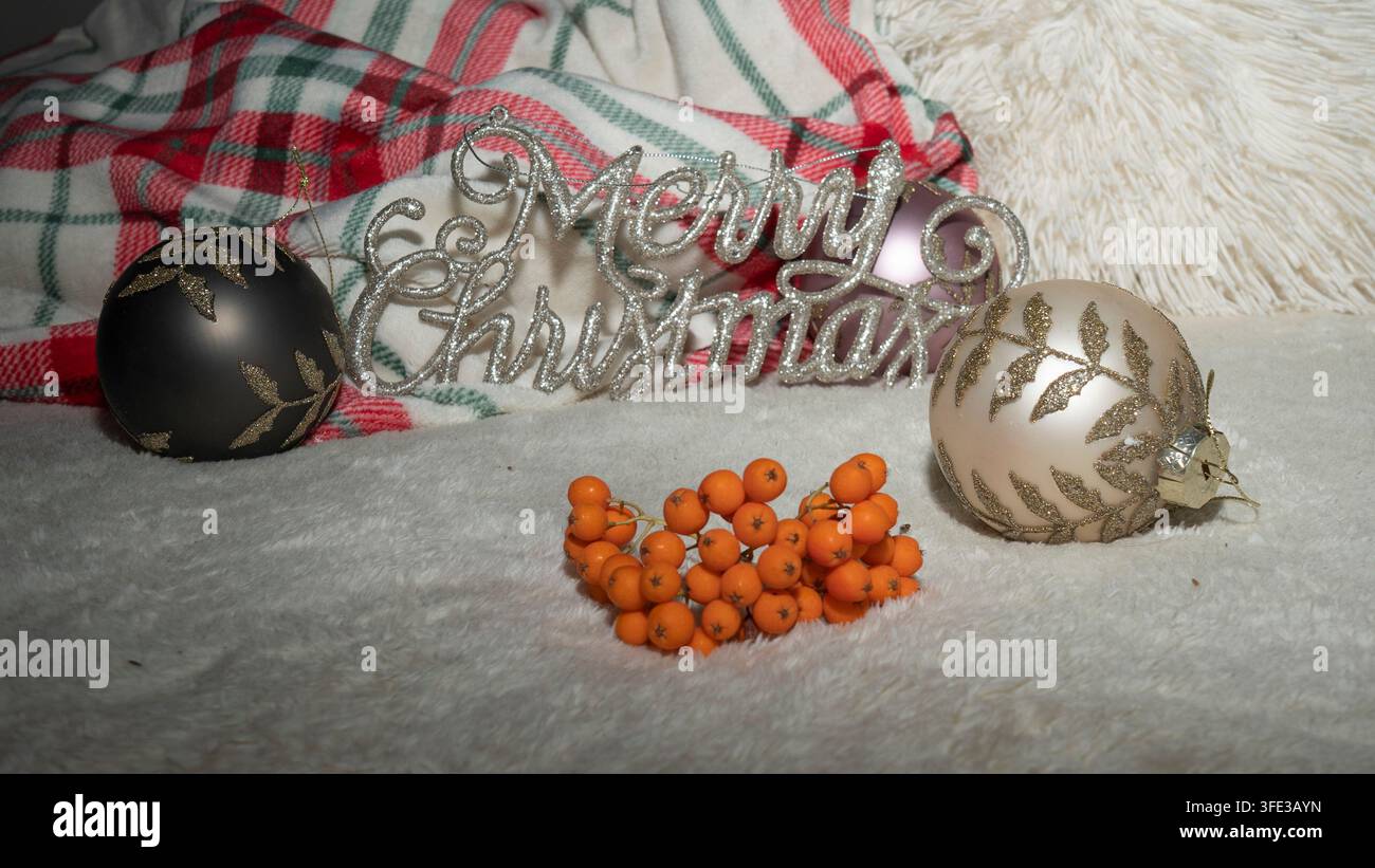 Insegna argentata scintillante con auguri di Buon Natale circondata da eleganti palline dell’albero e bacche di sorbo arancioni brillanti su una coperta di peluche bianca, morbido tartan a quadri rossi Foto Stock