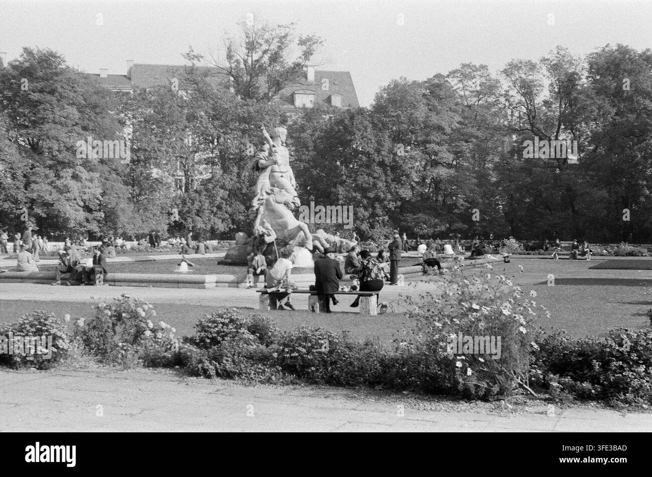 Fotografia in bianco e nero della metà del XX secolo di persone che si rilassano in un parco cittadino europeo intorno a una statua equestre ornata, circondata da alberi rigogliosi Foto Stock