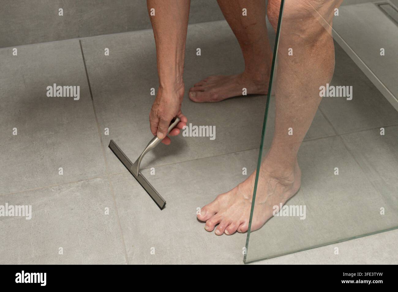 Persona che pulisce le piastrelle moderne del pavimento della doccia con un tergitore metallico Foto Stock