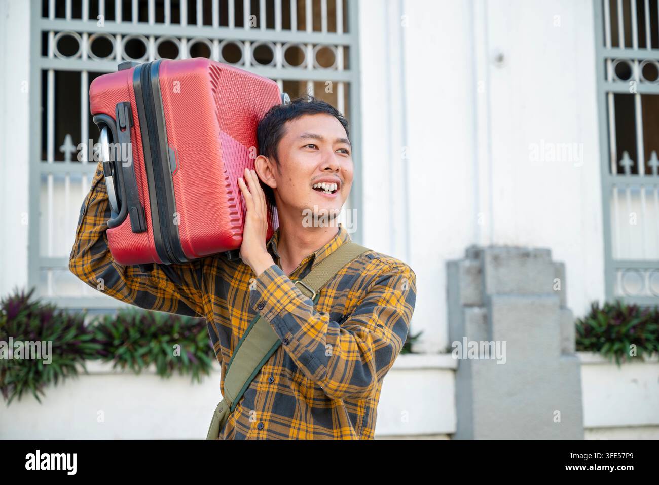 Un felice viaggiatore maschile indonesiano del sud-est asiatico in una camicia a quadri porta giocosamente una valigia rossa sulla spalla mentre è all'aperto davanti a Foto Stock