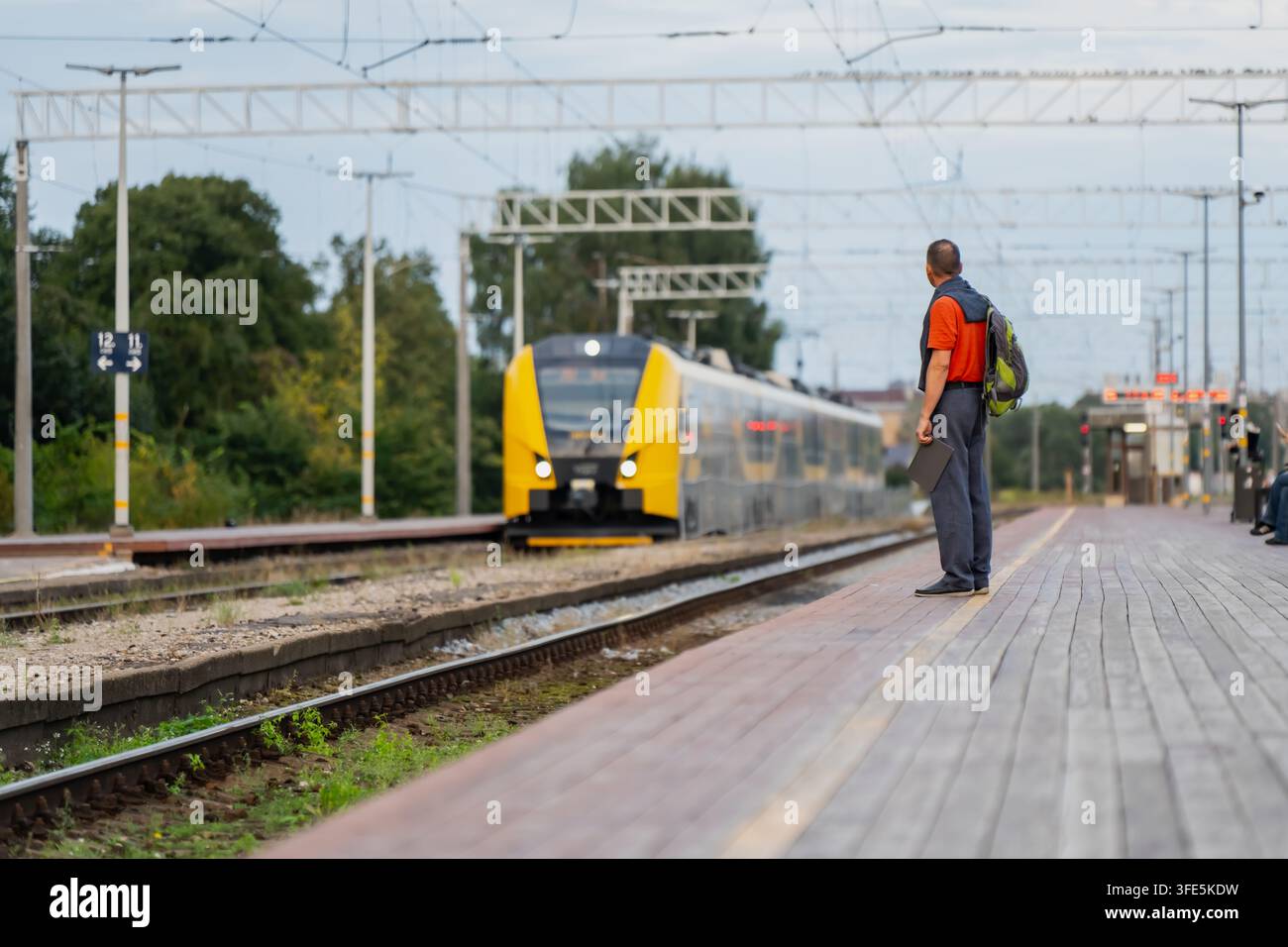 Le fermate ferroviarie regionali dipendono da un servizio poco frequente ma affidabile, un singolo passeggero in attesa da solo mentre un treno giallo si avvicina sotto una rete di linee aeree Foto Stock