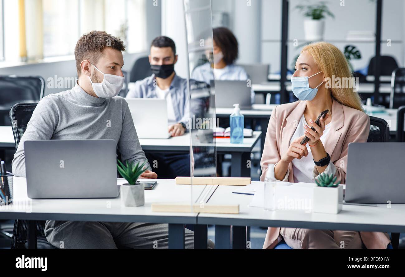 Uomo e donna mantengono la distanza sociale al lavoro. I dipendenti discutono del lavoro seduti a un tavolo con laptop, smartphone e vetro protettivo all'interno Foto Stock