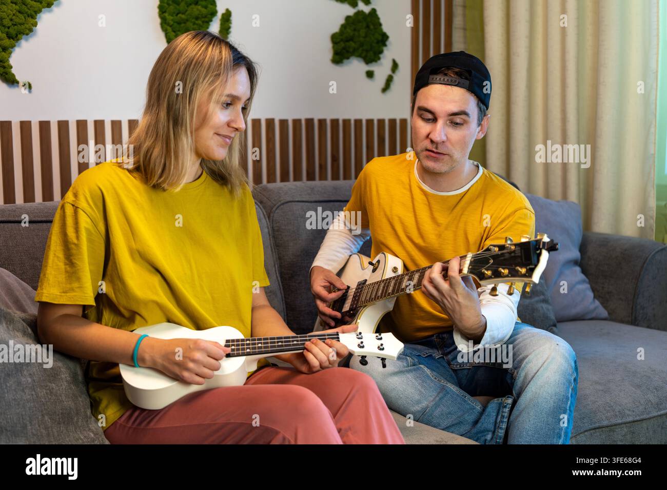 Coppia seduta su un divano a casa, che suona e prova musica sulle chitarre. Imparare a suonare, divertirsi insieme e godersi una lezione di musica. Foto Stock