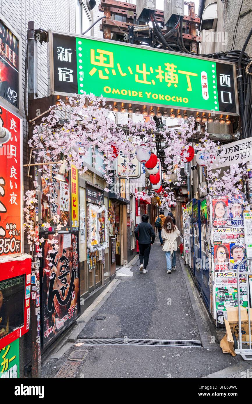 Persone che camminano attraverso il vivace vicolo Omoide Yokocho fiancheggiato da negozi, bar izakaya e ristoranti. Insegna sopra la testa e fiori di ciliegio di plastica Foto Stock