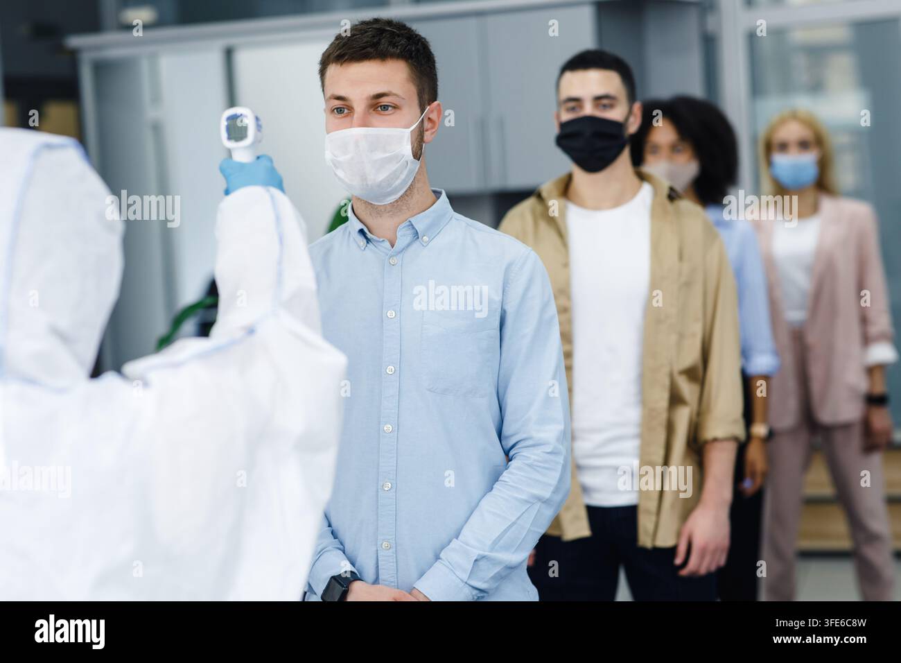 L'uomo in tuta protettiva misura la temperatura dei lavoratori in maschere protettive all'interno dell'ufficio, bloccando la diffusione di covid-19 Foto Stock