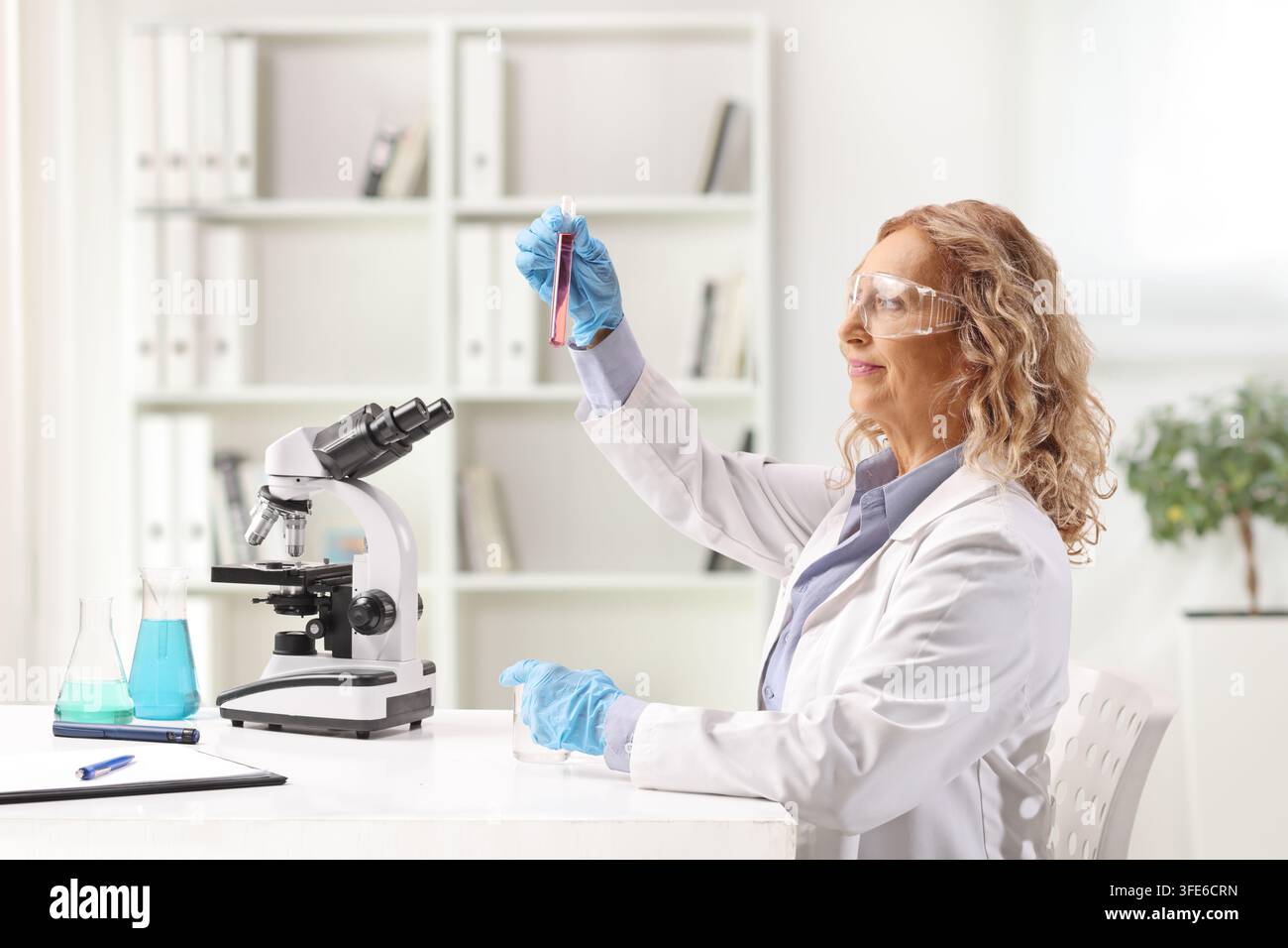 Scienziata donna che guarda una provetta e siede davanti a un microscopio su una scrivania Foto Stock