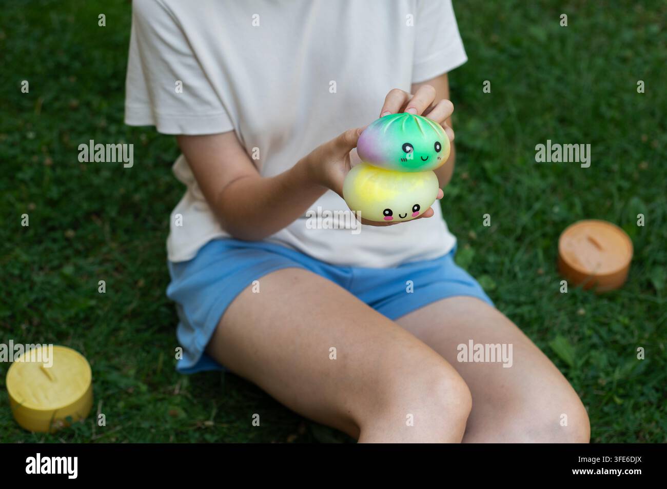 Bambino seduto sull'erba del prato in un giardino verde che gioca con squishies di dumpling kawaii colorati e contenitori per vapore. Foto Stock