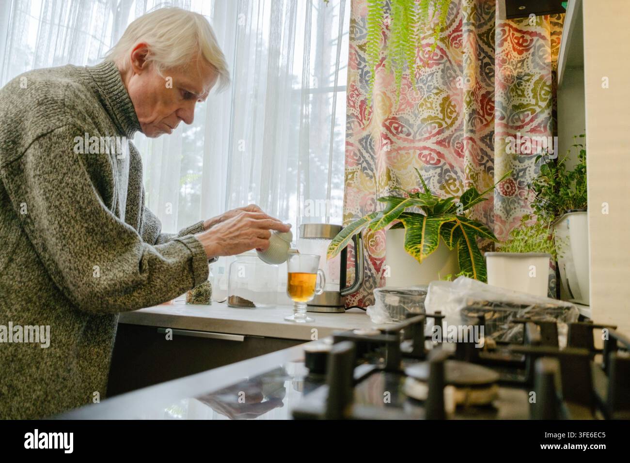 Un anziano si versa il tè da solo in cucina, trasmettendo un senso di solitudine. L'immagine riflette i momenti tranquilli della vita da soli nella vecchiaia. Nonno in una cucina vuota. Foto Stock