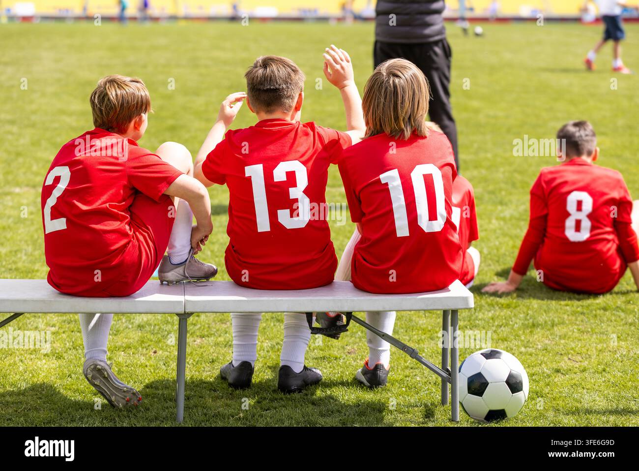 Giocatori di calcio giovanile in maglie rosse numerate seduti sulla panchina laterale e sull'erba a guardare la partita - Gestione della squadra di calcio junior Foto Stock