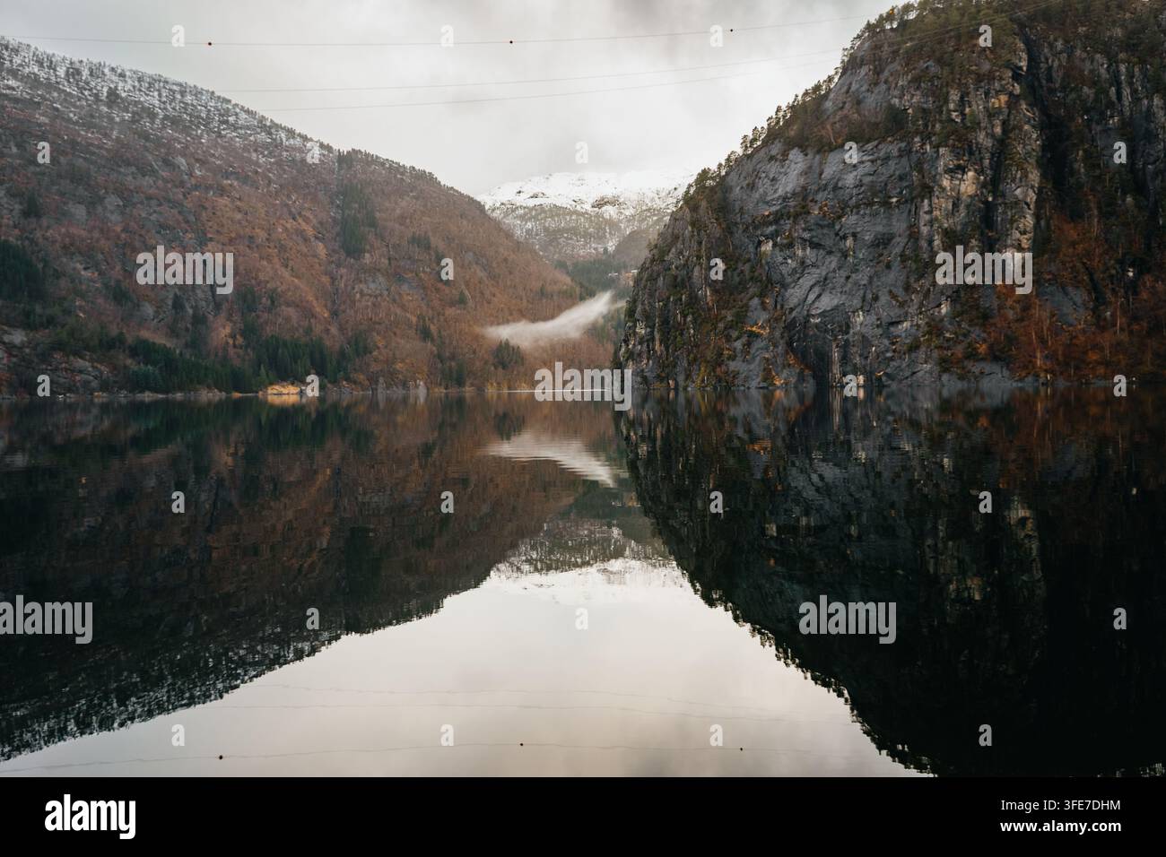 Nebbia bassa e nuvole sottili si sospendono tra ripide pendici boschive e scogliere rocciose scure, mentre l'acqua calma e scura crea un riflesso specchiato quasi perfetto Foto Stock