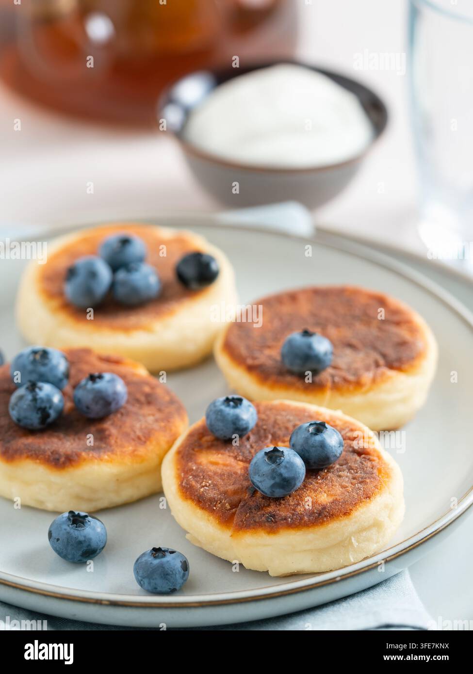 Deliziosi pancake casalinghi al curdo serviti con mirtilli su un piatto. Colazione sana del mattino con panna acida, idea di ricetta dolce. Sfondo luminoso e pulito con spazio di copia per il design del banner Foto Stock