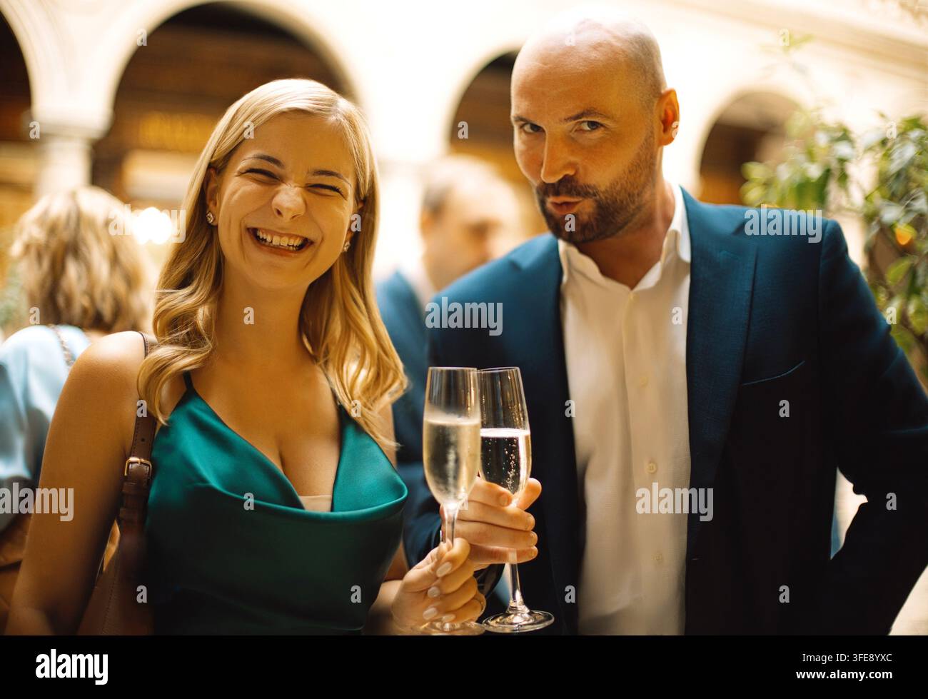 Donna caucasica in abito verde e uomo in completo che posano con bicchieri di champagne a un evento sociale, circondati da un'elegante architettura interna e da ospiti Foto Stock