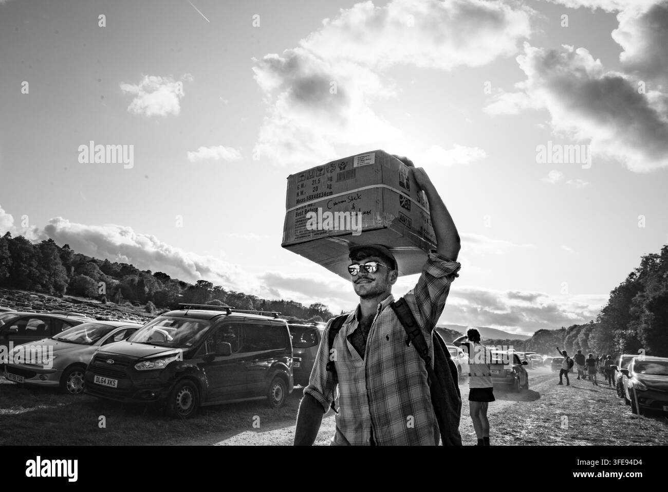 ARRIVO AL FESTIVAL CON SCATOLONI, REPORTAGE, GREEN MAN FESTIVAL 2019, BRECON, WALES, UK, 2019: Un giovane sorridente con occhiali da sole e cappellino porta grandi scatoloni sulla testa attraverso un parcheggio del festival illuminato dal sole in bianco e nero. Foto: Rob Watkins. INFO: Il Green Man Festival è iniziato nel 2003 in Galles. Si tiene annualmente nel terzo fine settimana di agosto nelle Brecon Beacons e celebra musica alternativa, arte, letteratura, scienza e cultura a misura di famiglia in uno splendido scenario montano. Foto Stock