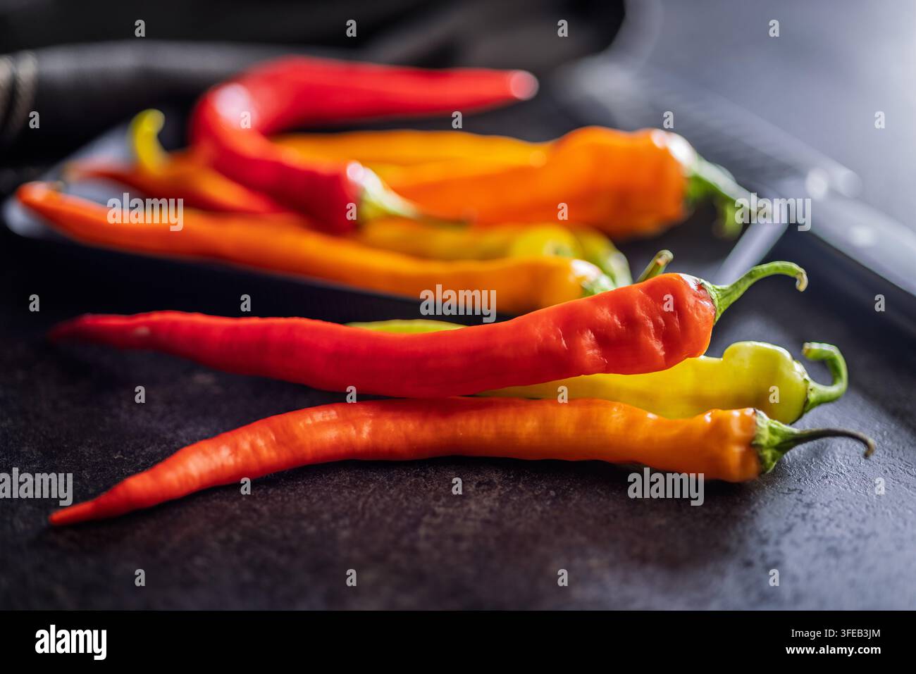 Lunghi peperoncini rossi, arancioni e verdi disposti su una superficie scura e ruvida; prodotti freschi e vibranti che evocano calore, spezia e preparazione culinaria rustica Foto Stock