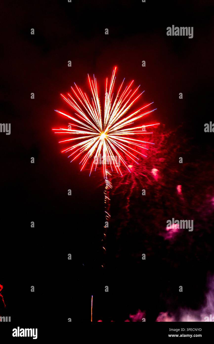 Colpi di fuoco d'artificio rossi vividi e dorati si estendono attraverso il cielo scuro della sera, irradiando luce a raggiera mentre il fumo arricciato si sposta sotto durante i festeggiamenti del quattro luglio Foto Stock
