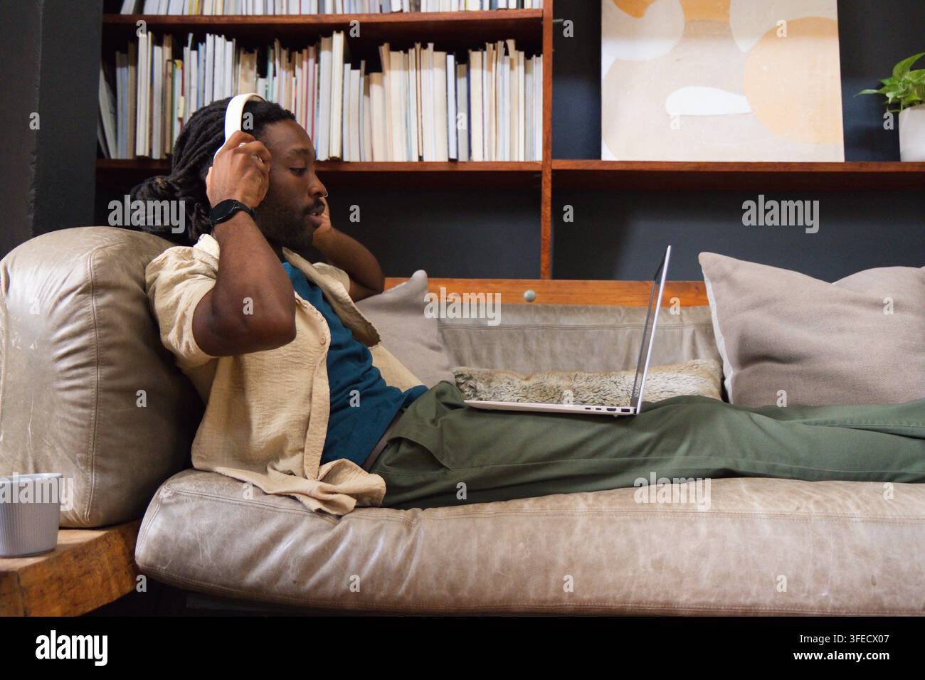 Uomo africano che si rilassa su un divano grigio a casa, aggiustando delle cuffie bianche mentre usa un laptop Foto Stock