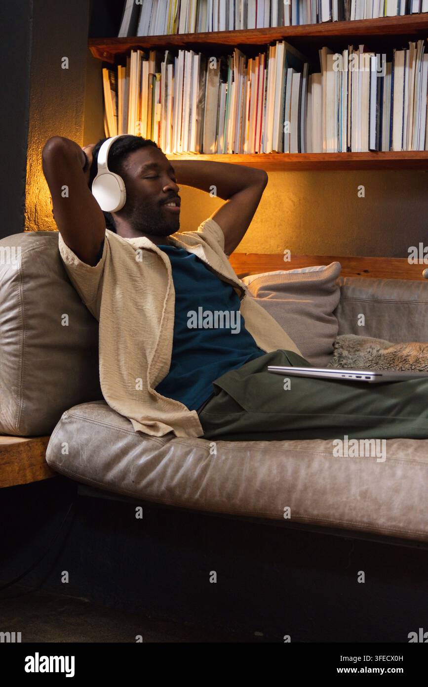 Uomo adulto afroamericano sdraiato sul divano a casa, con cuffie bianche e che usa un laptop Foto Stock