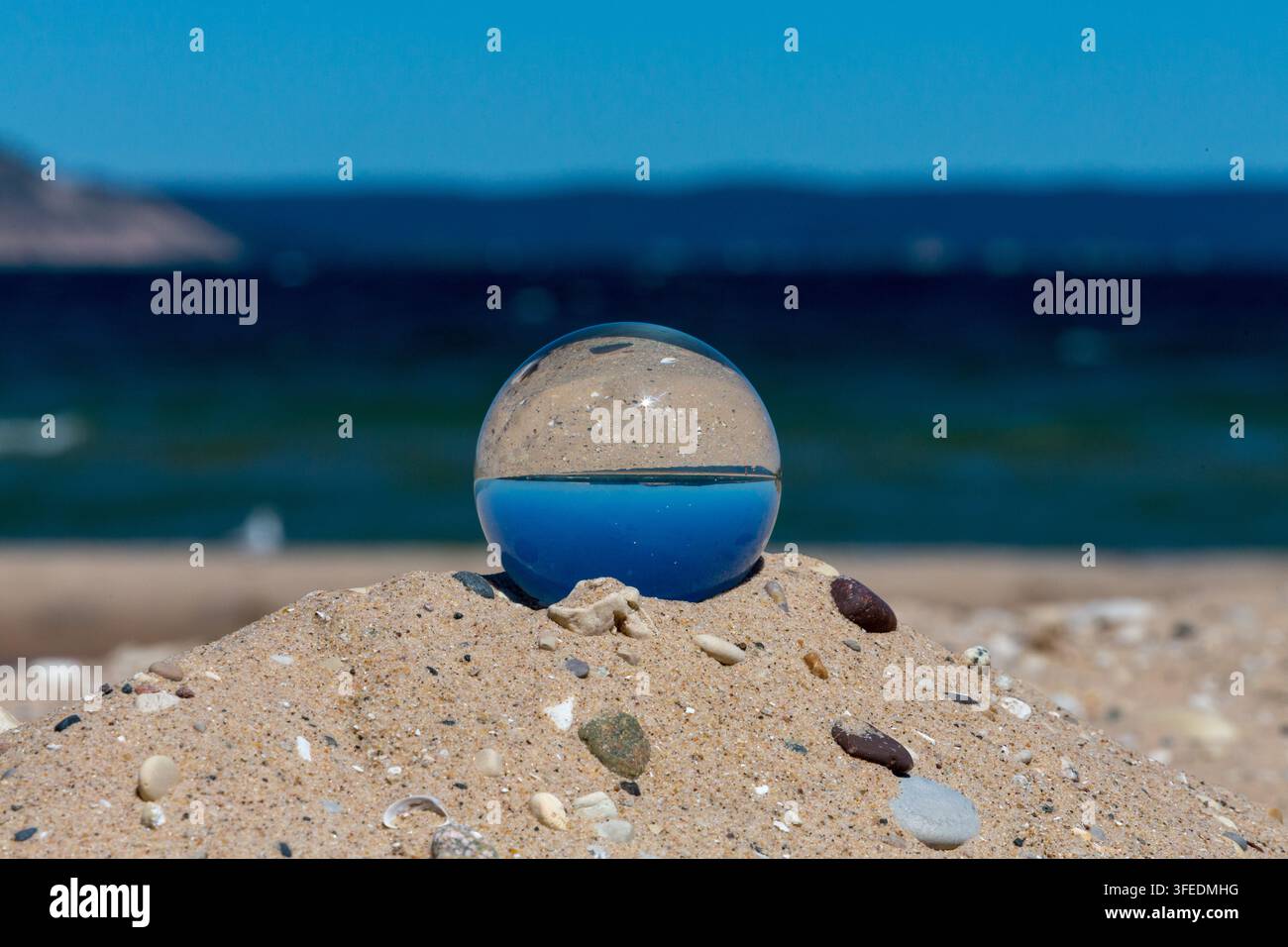 Lensball trasparente appoggiato su un cumulo di sabbia su una spiaggia soleggiata, che riflette le acque oceaniche invertite e il cielo azzurro nella fotografia a rifrazione creativa. Foto Stock