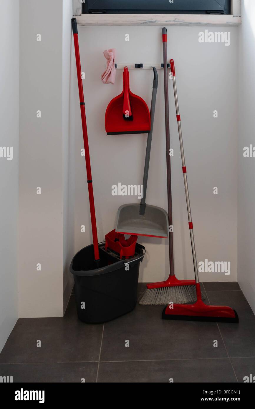 Scopa rossa, scopa con manico, paletta per la polvere e secchio per lo straccio stanno in un armadio bianco stretto con una piccola finestra sopra. Faccende domestiche e organizzazione della casa Foto Stock