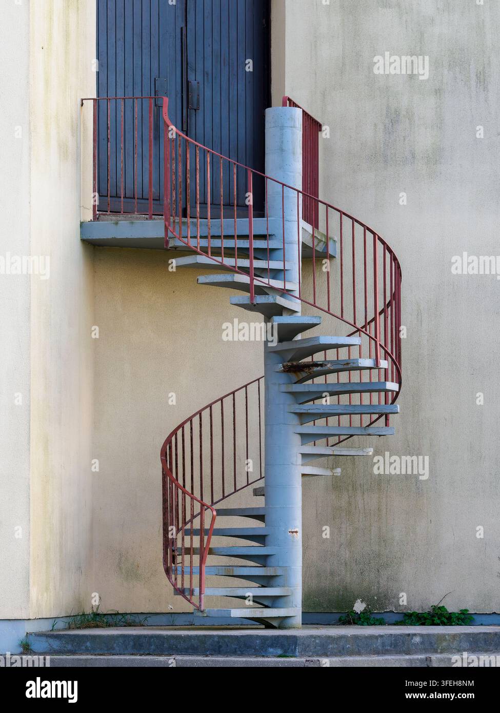 Scalinata a chiocciola in cemento attaccata all'esterno di un edificio, con una ringhiera in metallo rosso arrugginita che conduce a una porta di legno chiusa Foto Stock
