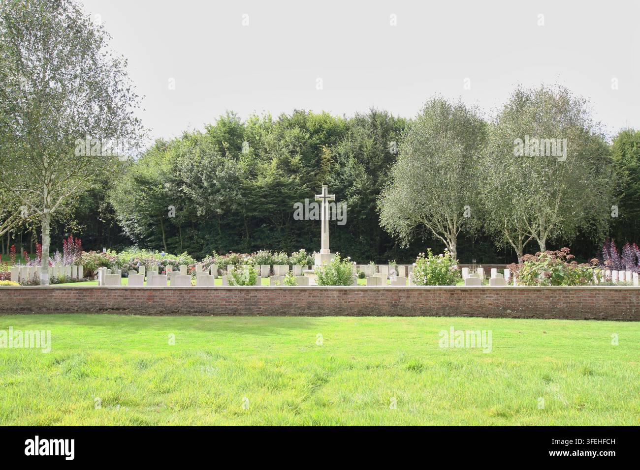 La croce bianca in pietra del Sacrificio si erge tra file di lapidi dietro un basso muro di contenimento in mattoni in un cimitero militare del Commonwealth in Belgio. Foto Stock