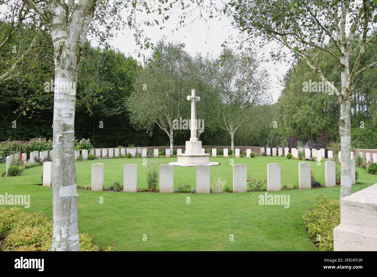 La croce bianca in pietra del Sacrificio si erge tra file di lapidi dietro un basso muro di contenimento in mattoni in un cimitero militare del Commonwealth in Belgio. Foto Stock