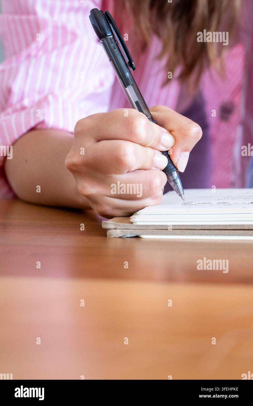 Primo piano di una persona che scrive in un quaderno con una penna nera su una scrivania di legno, prendendo appunti e studiando con concentrazione mentre indossa una camicia a righe rosa Foto Stock