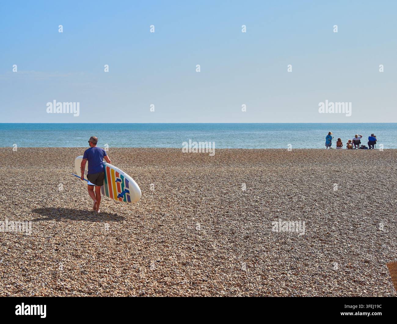Surfer che trasporta una tavola da remo attraverso una spiaggia di ciottoli verso il mare calmo in una giornata estiva soleggiata Foto Stock