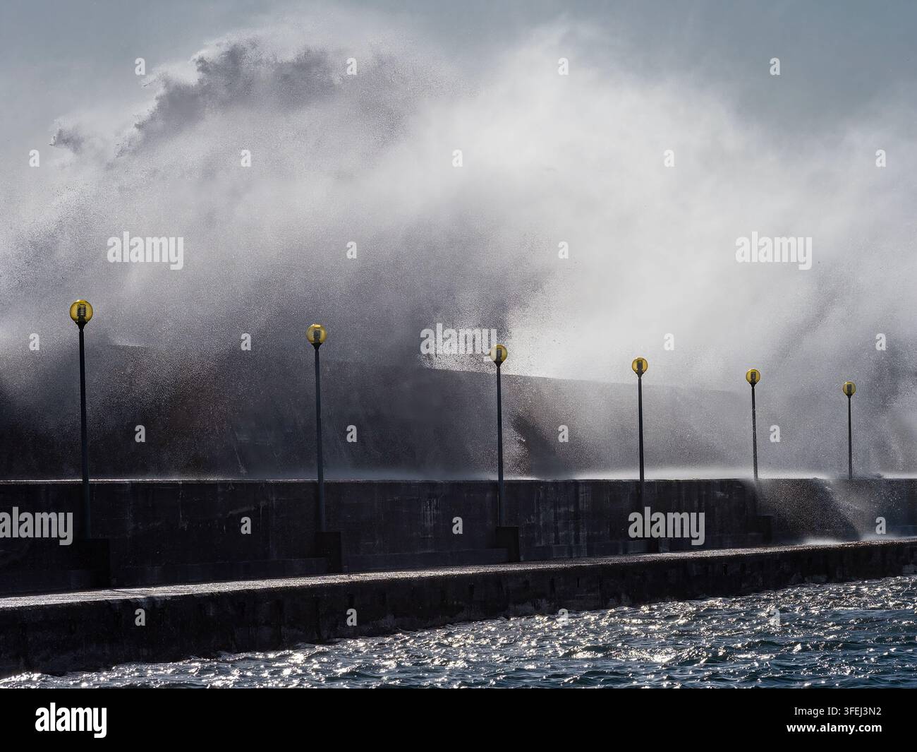 Onde gigantesche che si infrangono su un muro di protezione in un porto con lampioni durante una grave tempesta costiera Foto Stock